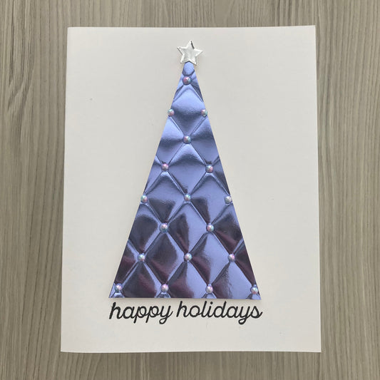 A handmade holiday greeting card featuring a metallic lavender quilted triangle tree with a silver star topper and the words 'happy holidays' in black script, displayed on a light wood background.