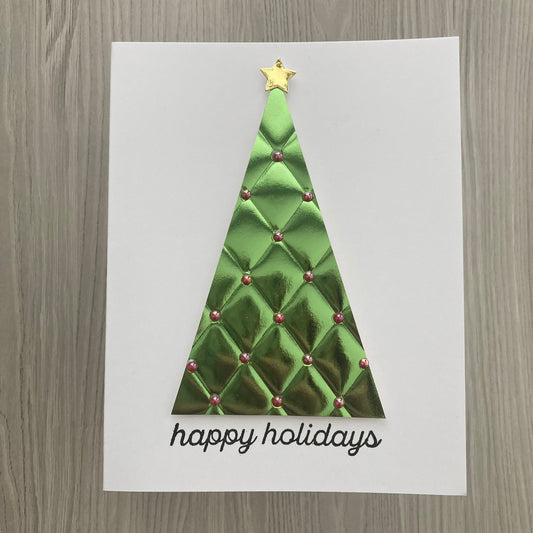 A white greeting card featuring a metallic green quilted Christmas tree with a gold star on top and the words 'happy holidays' in black script at the bottom, displayed on a gray wood-grain surface.
