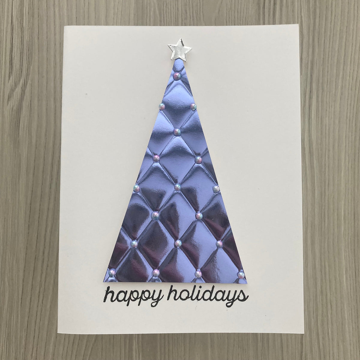 A handmade holiday greeting card featuring a metallic lavender quilted triangle tree with a silver star topper and the words 'happy holidays' in black script, displayed on a light wood background.