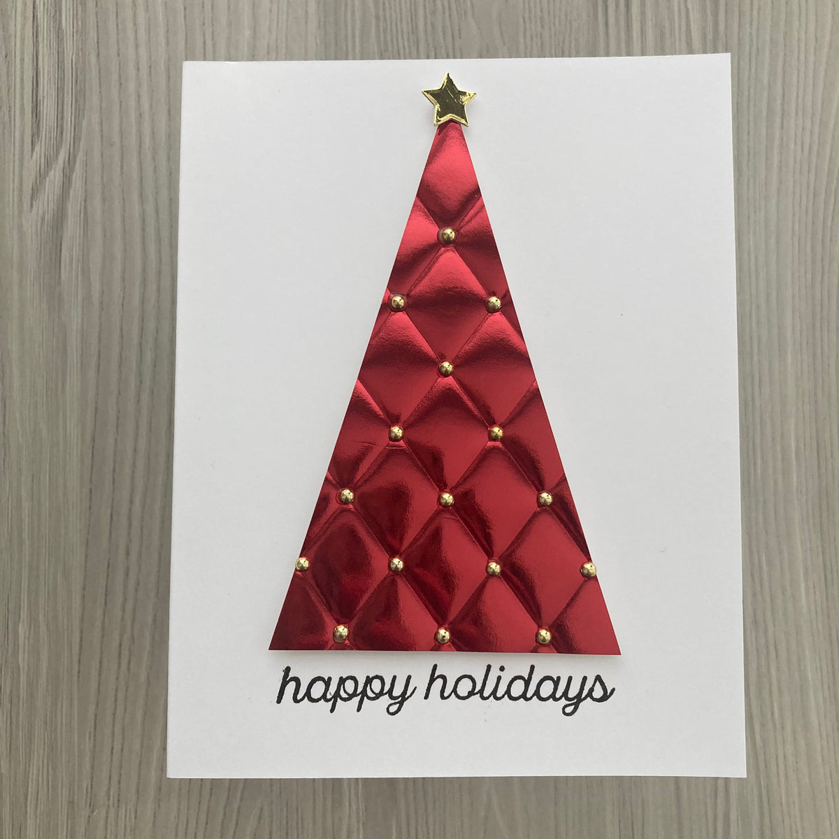 A square white greeting card featuring a red quilted Christmas tree design with small gold studs and a gold star on top, plus the words 'happy holidays' in black script at the bottom, photographed on a gray wood-textured surface.