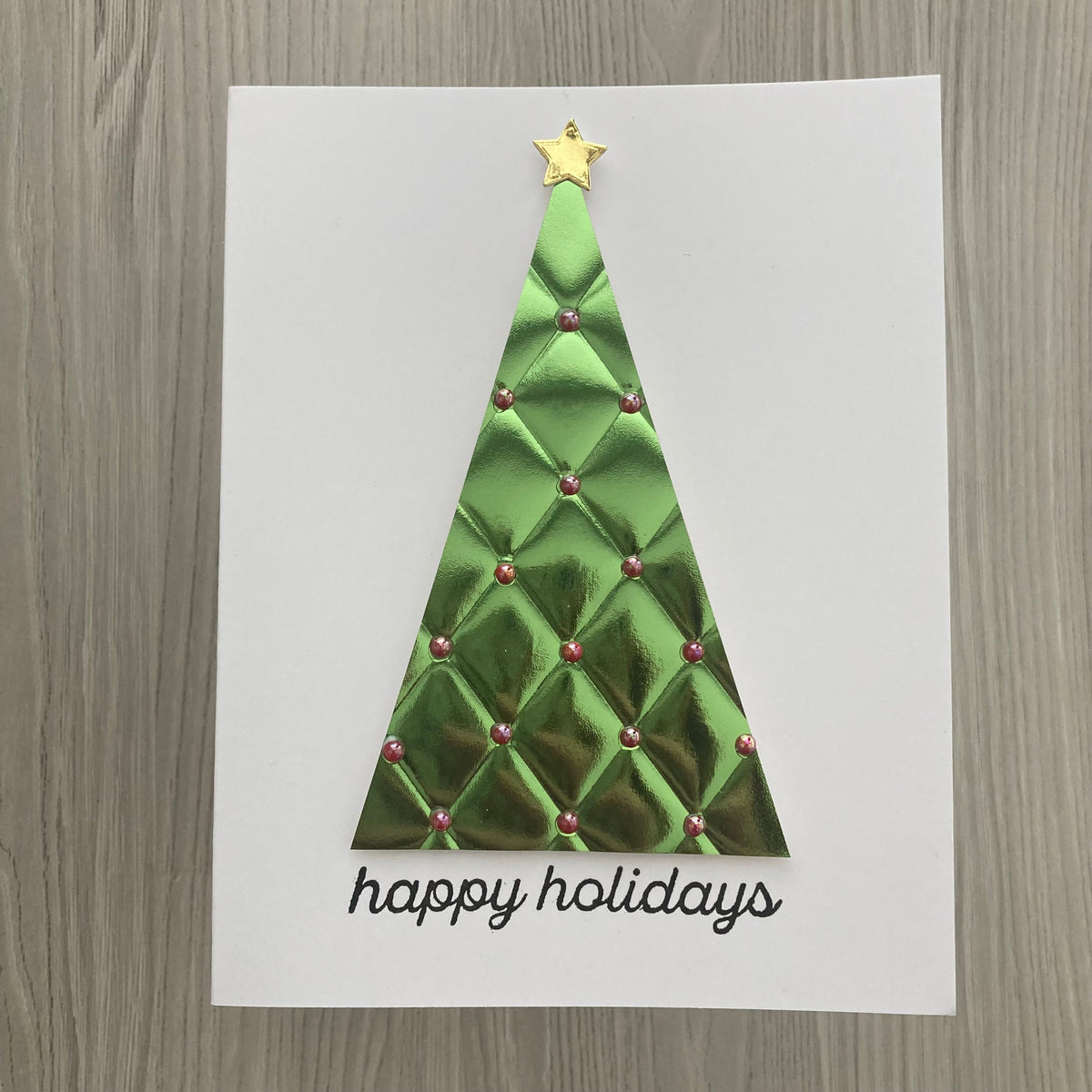 A white greeting card featuring a metallic green quilted Christmas tree with a gold star on top and the words 'happy holidays' in black script at the bottom, displayed on a gray wood-grain surface.