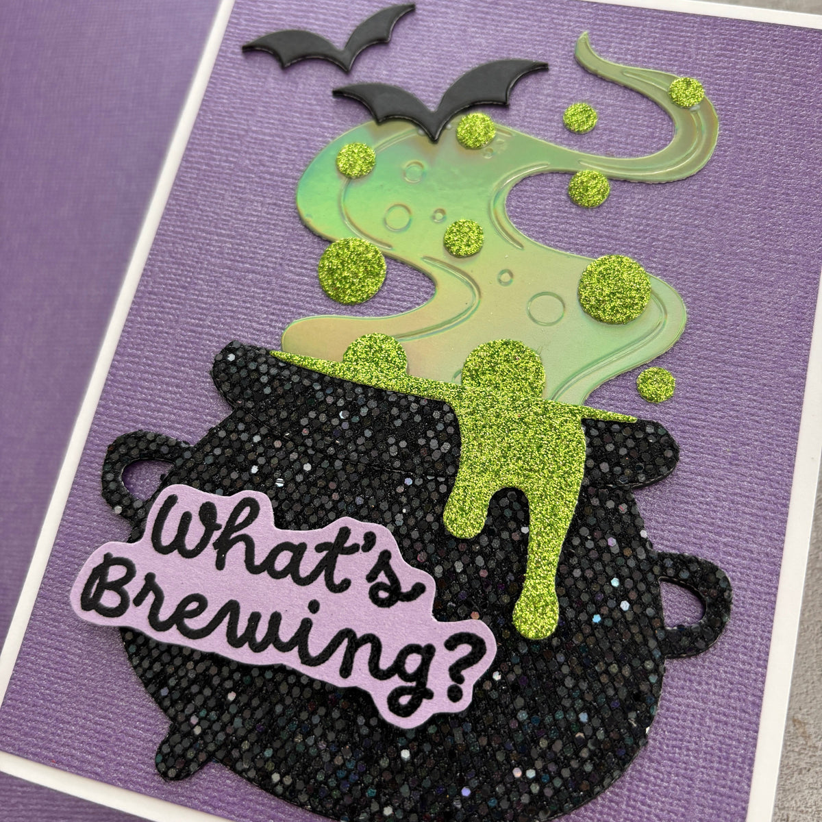 handmade Halloween card featuring a black glitter cauldron with the phrase 'What's Brewing?' in black script on a lavender die-cut sticker