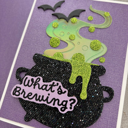 handmade Halloween card featuring a black glitter cauldron with the phrase 'What's Brewing?' in black script on a lavender die-cut sticker