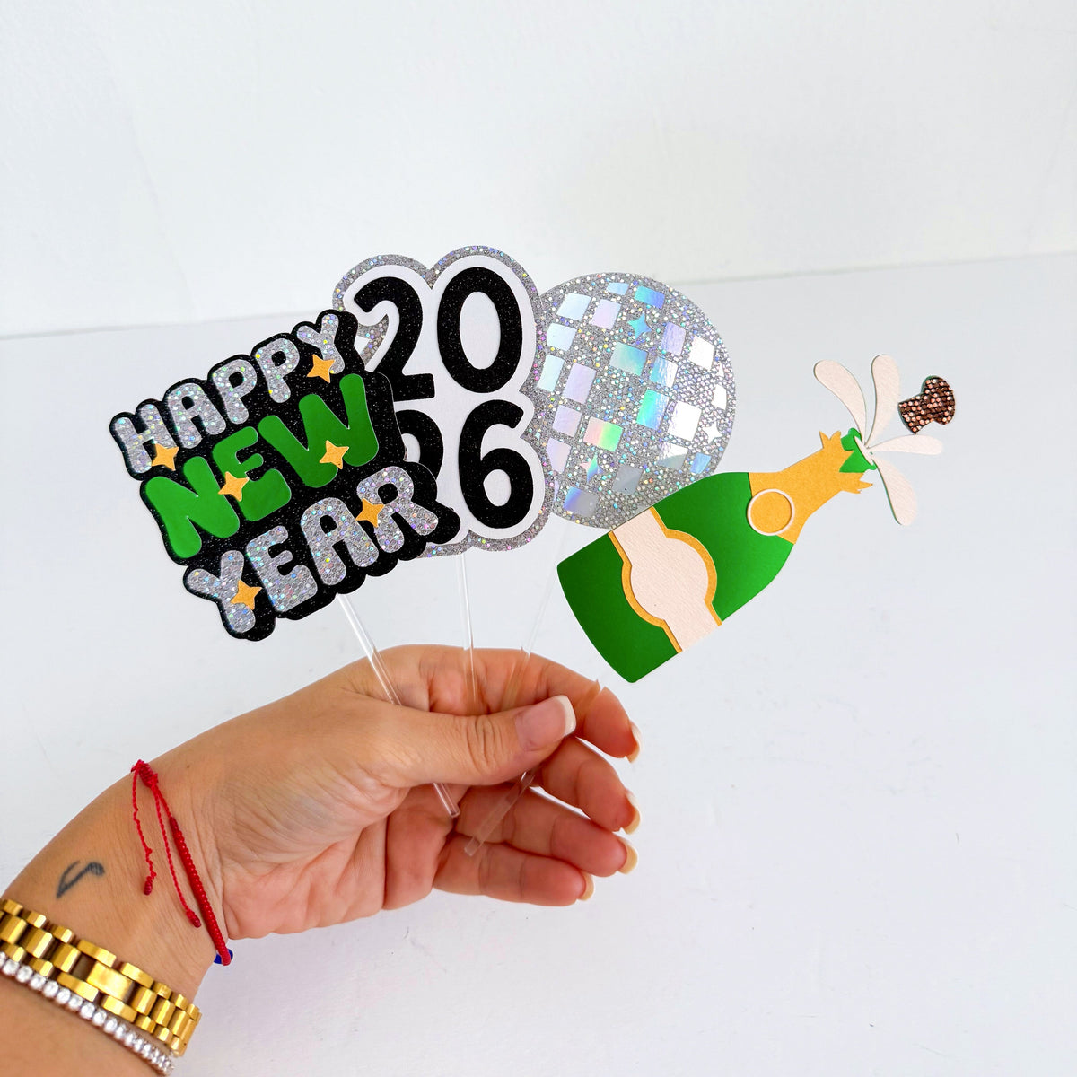 Hand holding Happy New Year 2026 cupcake topper decoration with a bottle of champagne on a white background