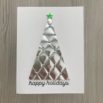 A white greeting card featuring a textured, diamond-quilted silver Christmas tree with a small green star on top and the words 'happy holidays' in black script below, displayed on a gray wood-grain surface.